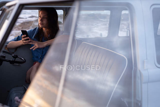 Вид збоку вдумливі кавказьких жінці за допомогою мобільного телефону в camper van на пляжі — стокове фото