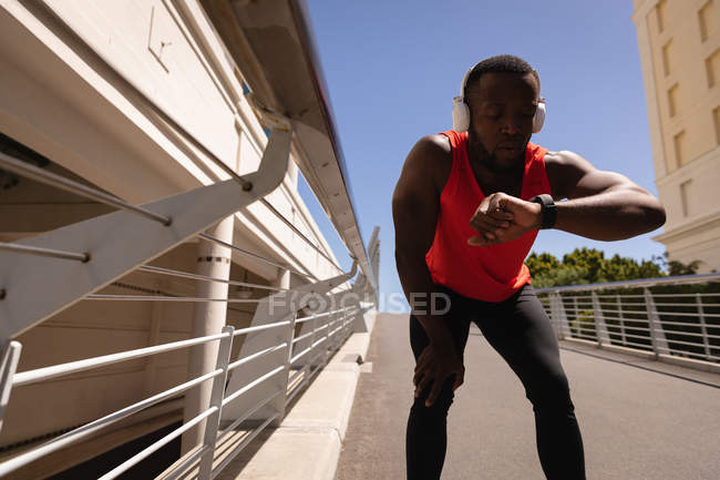 Frontansicht eines jungen afrikanisch-amerikanischen fitten Mannes, der auf einer Brücke entspannt auf eine Smart Watch blickt — Stockfoto