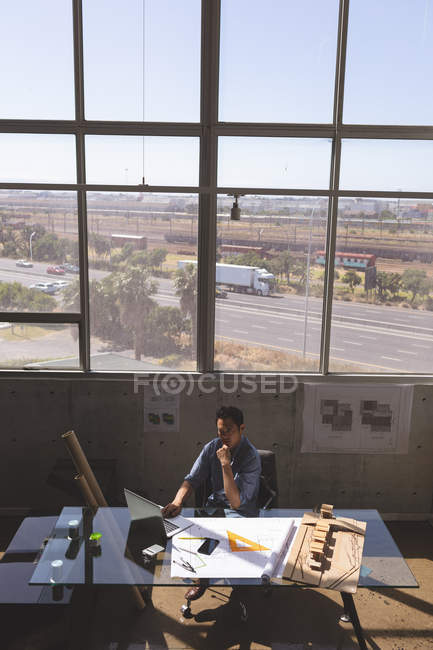 Vista de ángulo alto del arquitecto asiático sentado en el escritorio y trabajando en el ordenador portátil en una oficina moderna - foto de stock