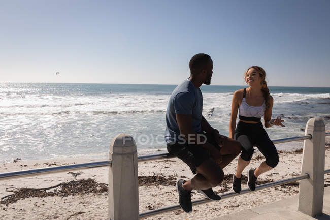 Seitenansicht eines jungen multiethnischen Paares, das an einem sonnigen Tag auf einem Geländer an der Strandpromenade sitzt — Stockfoto