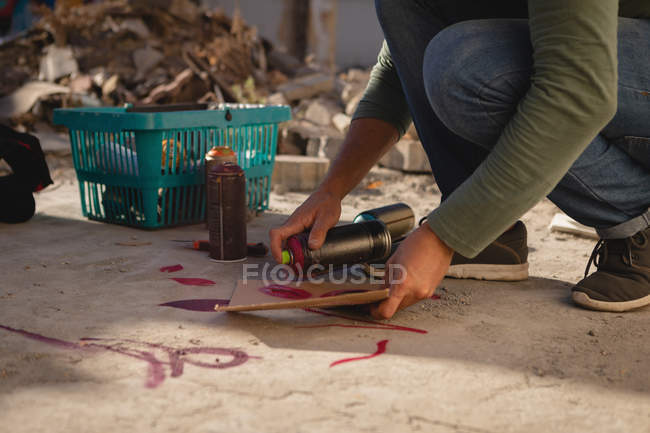 Low section of young Caucasian graffiti artist spray painting on card board at alley — Stock Photo