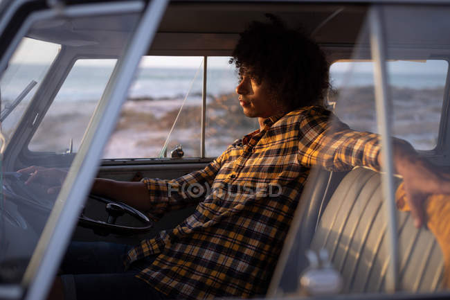 Vista lateral do homem de corrida mista relaxado dirigindo uma van campista contra o oceano no fundo, enquanto o pôr do sol — Fotografia de Stock