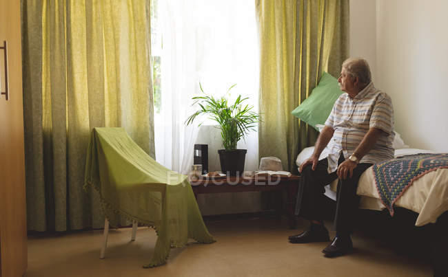 Вид сбоку на грустного пациента старшего кавказца, выглядывающего из окна, сидящего в одиночестве на кровати дома престарелых — стоковое фото