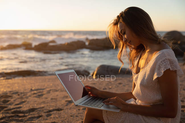 Vista lateral de la mujer tranquila usando el ordenador portátil en la playa al atardecer - foto de stock