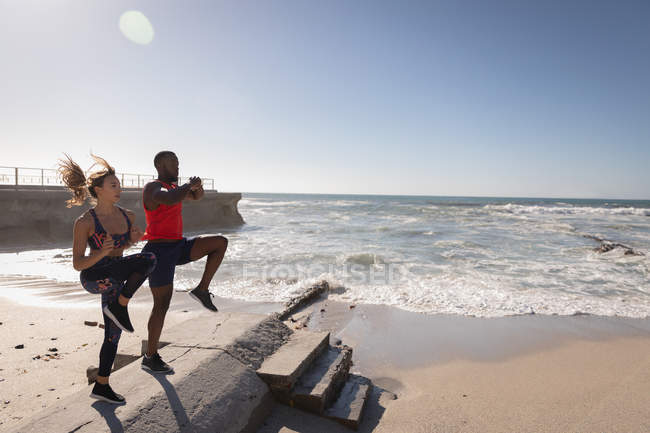 Vista lateral de una joven pareja multiétnica haciendo footing en la playa - foto de stock