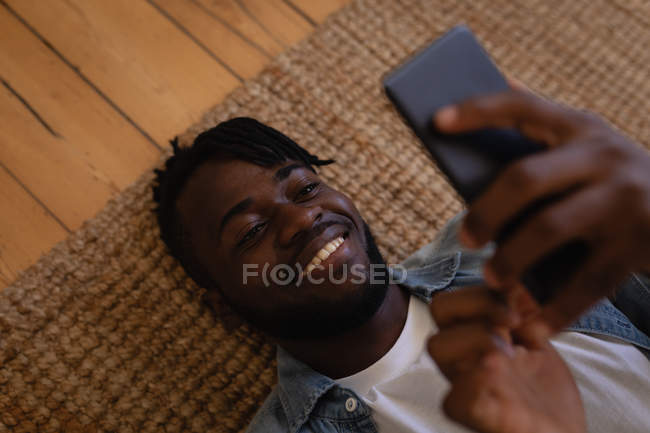 Vista de alto ângulo de homem afro-americano bonito usando telefone celular enquanto deitado no chão em casa. Ele está sorrindo. — Fotografia de Stock