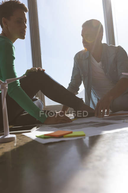Vue de face des architectes caucasiens assis sur le sol et discutant sur le plan dans le bureau. Ils sont assis à côté de la fenêtre et le soleil brille . — Photo de stock