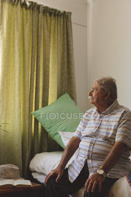 Side view of senior Caucasian man looking outside the window while sitting alone on nursing home bed — Stock Photo