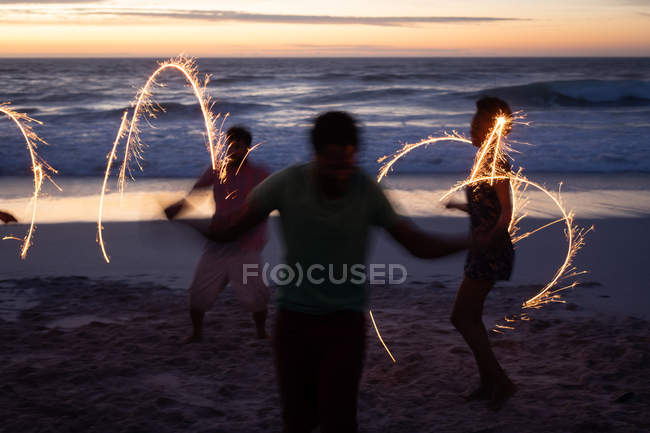 Vue arrière d'un groupe d'amis multiethniques appréciant sur la plage avec des étincelles au premier plan contre l'océan au premier plan — Photo de stock
