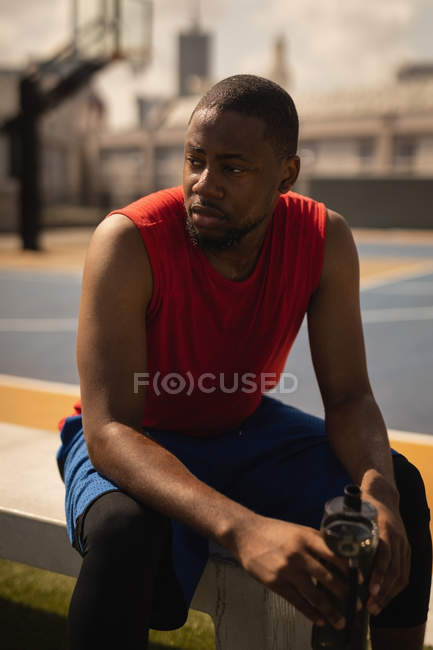 Vista frontal del jugador de baloncesto afroamericano en camiseta sin mangas relajándose en la cancha de baloncesto mientras tiene una botella de agua en sus manos - foto de stock