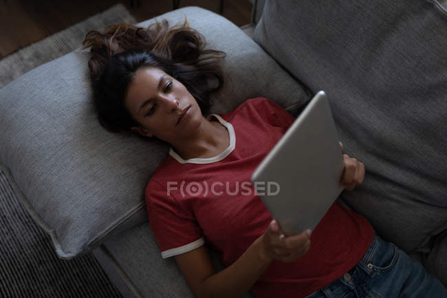 Високий кут зору красивої змішаної раси жінки, що використовує цифровий планшет, спираючись на диван вдома — стокове фото