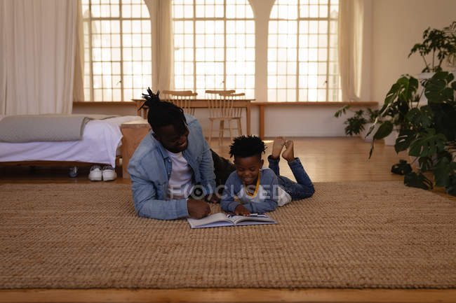 Front view of happy African-American father and son reading book while lying on floor at home — Stock Photo