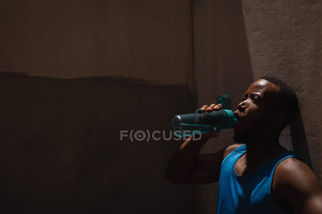 Side view of young African-American fit man drinking water while standing beside the wall on a sunny day — Stock Photo