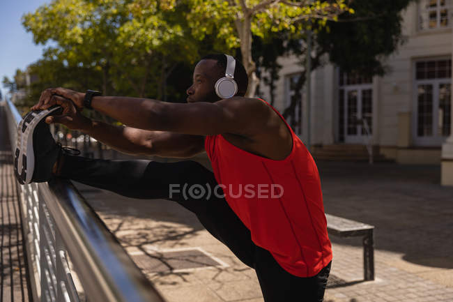 Vue latérale du jeune homme en forme afro-américain avec casque étirant la jambe contre la rampe sur la promenade latérale — Photo de stock