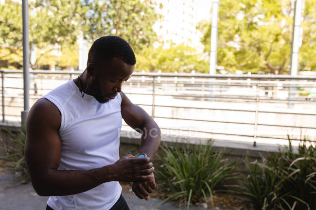 Vista laterale del giovane afro-americano in forma uomo guardando orologio intelligente sulla strada — Foto stock