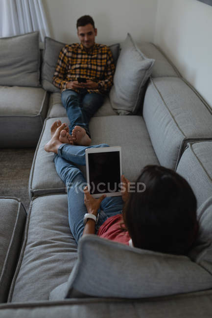 Heureux couple multiethnique utilisant une tablette numérique et un téléphone portable sur le canapé à la maison — Photo de stock