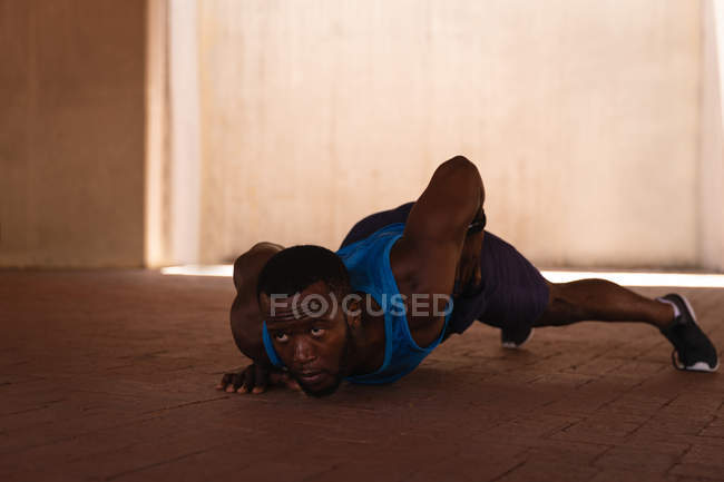 Front view of young African-American fit man doing one-arm push-up exercise under the bridge on a sunny day — Stock Photo