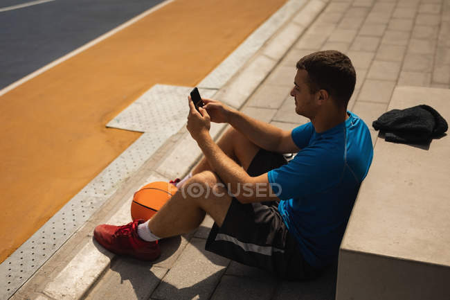 Vista de ángulo alto del joven jugador de baloncesto caucásico utilizando el teléfono móvil mientras se relaja en la cancha de baloncesto - foto de stock