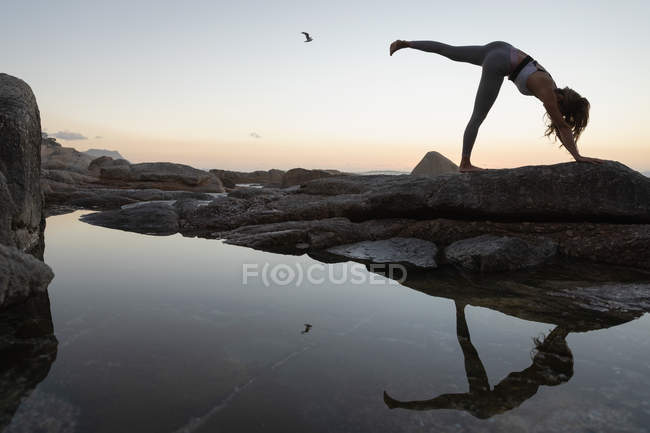 Low angle view of woman doing yoga on rock on the beach at sunset — Stock Photo