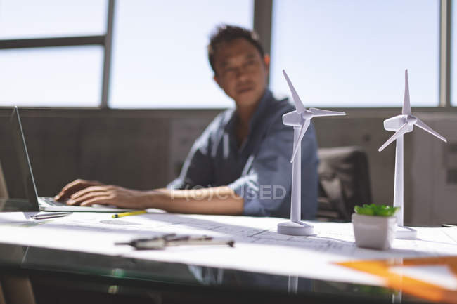 Vista laterale dell'architetto asiatico di sesso maschile che lavora su laptop alla scrivania in un ufficio moderno, turbina eolica in primo piano — Foto stock