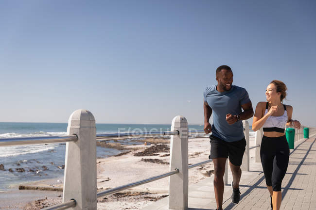 Вид на молодую многоэтническую пару, бегущую по набережной рядом с пляжем в солнечный день — стоковое фото