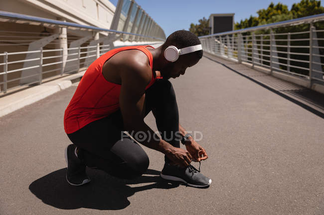 Вид збоку молодих афро-американських fit людина, прив'язавши його взуття мережива при crouching на тротуарі на сонячний день — стокове фото