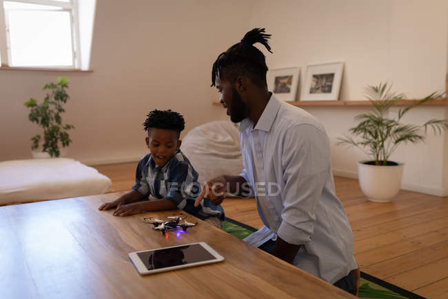 Vista lateral del apuesto padre e hijo afroamericanos jugando con un dron en la mesa en casa - foto de stock