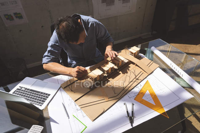 Vista de ángulo alto del arquitecto asiático masculino que trabaja en el modelo de edificio de oficinas con su computadora portátil y herramientas en una oficina moderna - foto de stock