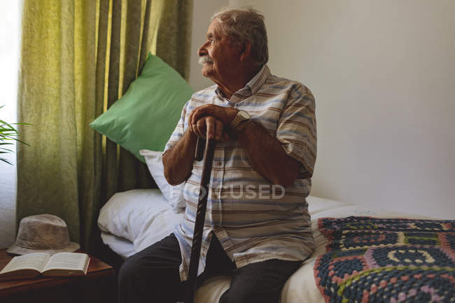 Вид спереди на старшего кавказца, смотрящего в окно, сидящего в одиночестве на кровати в доме престарелых — стоковое фото