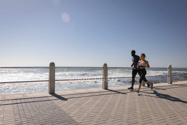 Seitenansicht eines jungen multiethnischen Paares beim Joggen auf dem Bürgersteig an der Strandpromenade — Stockfoto