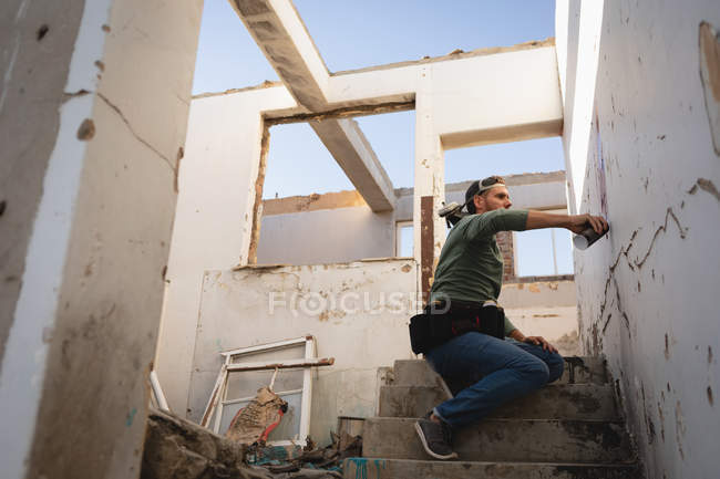 Niedrigwinkel-Ansicht junger kaukasischer Graffiti-Künstler sprüht Malerei auf verwitterte Wand, während er auf Treppen sitzt — Stockfoto