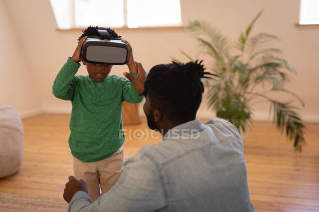 Vue arrière du père et du fils afro-américains interagissant les uns avec les autres tout en utilisant un casque de réalité virtuelle à la maison — Photo de stock