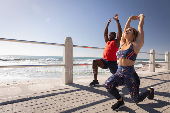 Vista laterale della giovane coppia multietnica che si allena sul marciapiede vicino alla spiaggia in una giornata di sole — Foto stock