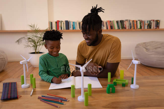 Vue de face du père afro-américain heureux aidant son fils à dessiner assis sur une chaise à la maison — Photo de stock