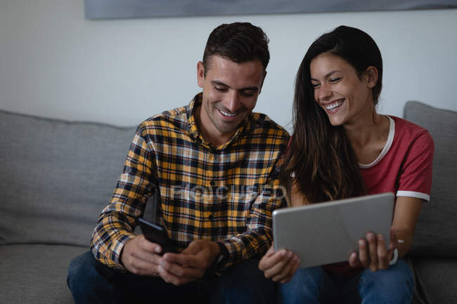 Вид на счастливую мультиэтническую пару с помощью мобильного телефона и цифрового планшета на диване дома — стоковое фото