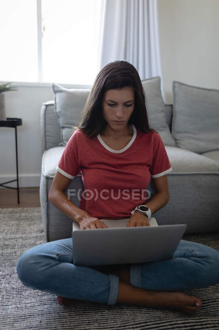 Vista frontal de la joven mujer de raza mixta utilizando el ordenador portátil sentado en el suelo en casa - foto de stock