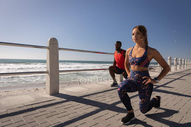 Vue de côté du jeune couple multi-ethnique faisant de l'exercice sur la chaussée près de la plage par une journée ensoleillée — Photo de stock