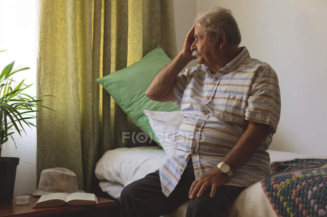 Vista lateral del hombre caucásico mayor mirando fuera de la ventana mientras está sentado solo y sosteniendo su cabeza en la cama del hogar de ancianos - foto de stock