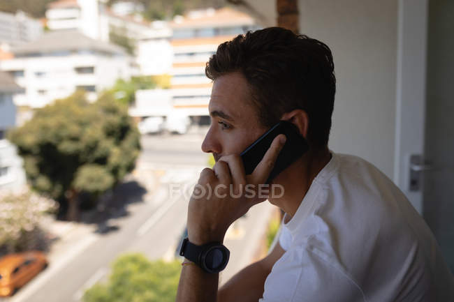 Вид сбоку на молодого кавказца, разговаривающего по мобильному телефону, стоя дома на балконе — стоковое фото