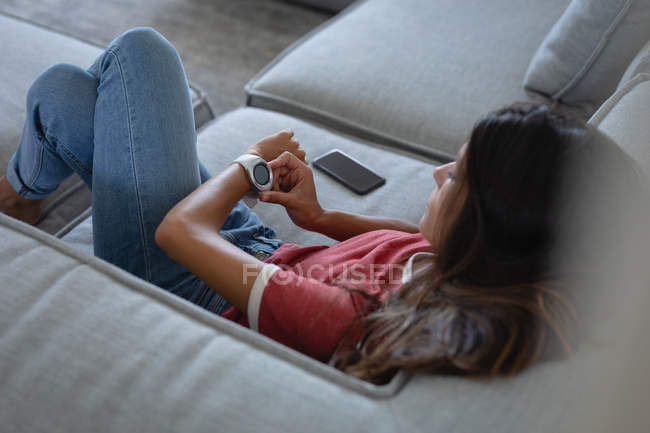 Високий кут зору змішаної раси жінки, використовуючи розумний годинник, спираючись на диван вдома — стокове фото