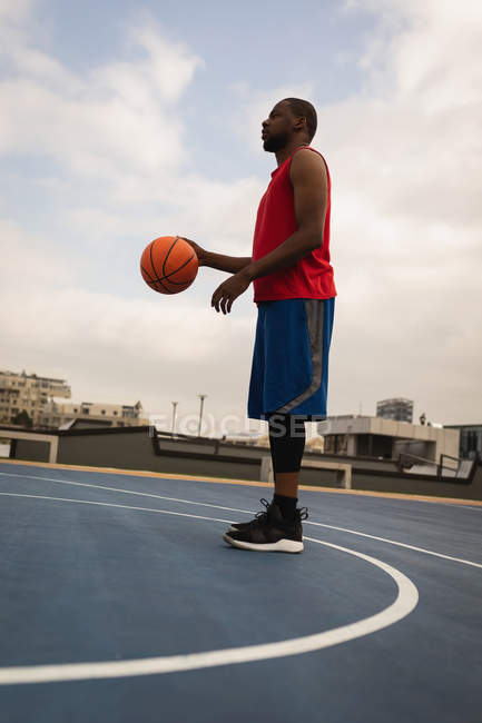Vista lateral del jugador de baloncesto afroamericano jugando baloncesto en la cancha de baloncesto contra la ciudad en segundo plano - foto de stock
