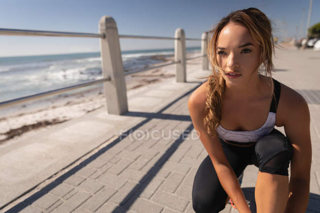 Vista frontal de la mujer atando zapato de encaje en el pavimento en la playa del paseo marítimo - foto de stock