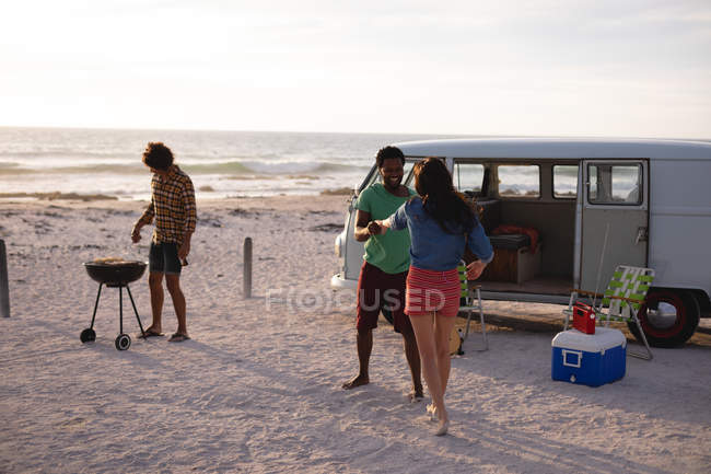 Vista frontal de amigos multi-étnicos dançando juntos na areia na praia, enquanto um outro fazendo um churrasco contra um oceano e praia no fundo — Fotografia de Stock