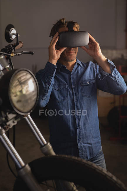 Vista frontal do mecânico de bicicletas masculino caucasiano usando fone de ouvido de realidade virtual na garagem — Fotografia de Stock