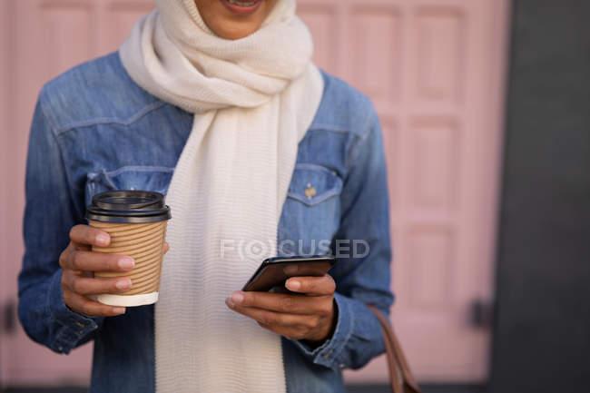 Средняя секция женщины, использующей мобильный телефон и держащей кофе, стоя перед дверью на улице — стоковое фото