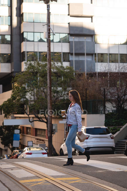 Vista lateral de la hermosa joven mujer caucásica con taza de café cruzando la calle en la ciudad - foto de stock