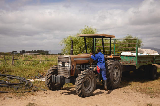 Вид сзади на афро-американского фермера в комбинезоне, садящегося на трактор на фермерской дороге — стоковое фото
