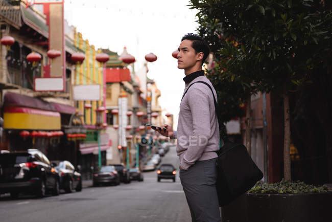 Vista lateral del hombre asiático reflexivo usando el teléfono móvil mientras está de pie en la calle - foto de stock