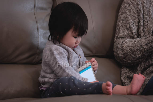 Vorderansicht eines kleinen süßen asiatischen Babys, das mit dem Handy spielt, während es zu Hause neben seiner Mutter auf dem Sofa sitzt — Stockfoto