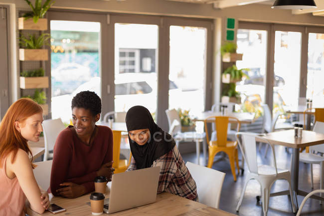 Seitenansicht von jungen Freundinnen gemischter Rassen, die miteinander interagieren, während sie Laptop in einem Café benutzen — Stockfoto